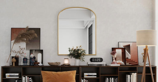 A golden arch mirror is mounted on 2 walnut storage cabinets with crafts and paintings on the countertops