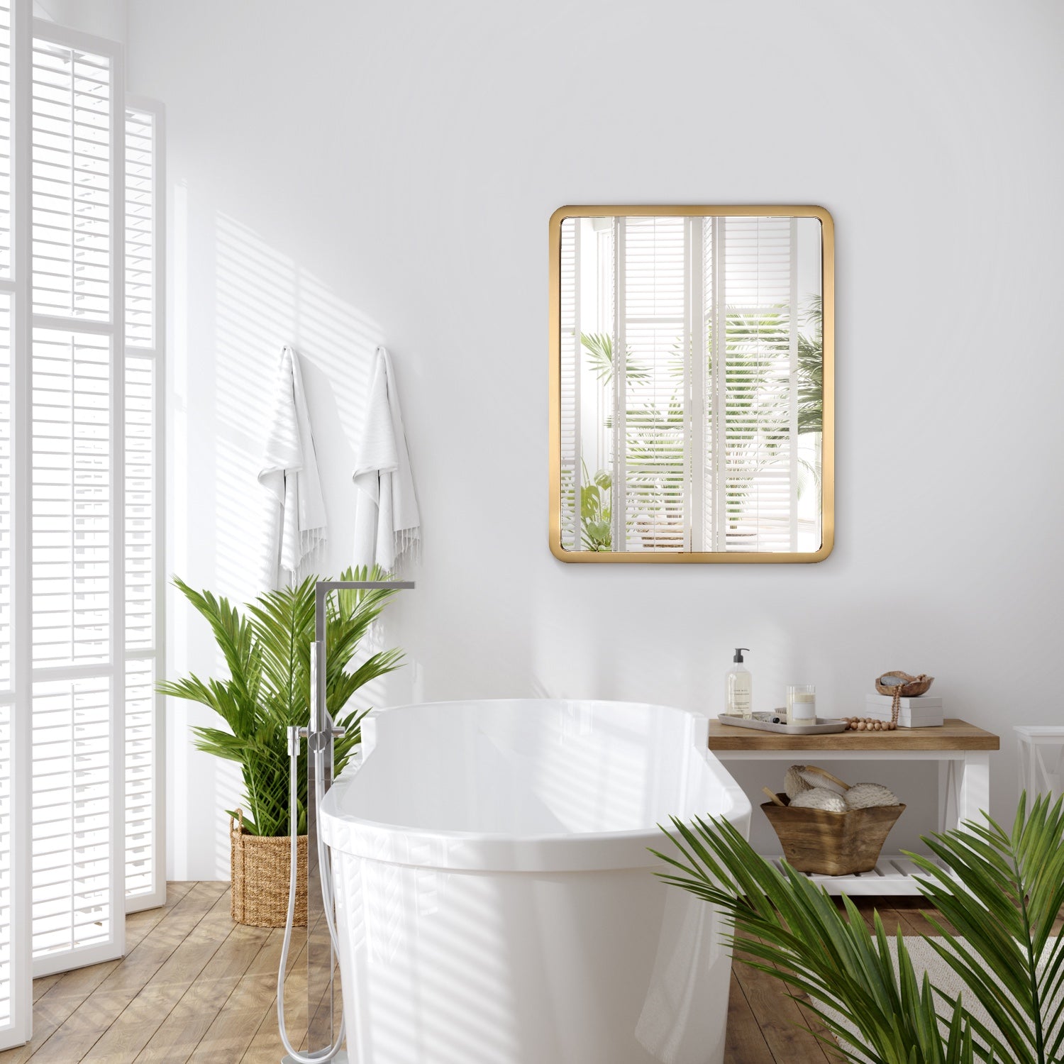 In a bathroom adorned with white shutters, a luxurious gold rectangle mirror reflects natural light above a white bathtub, with two white towels hanging nearby and greenery around the floor, enhancing relaxation